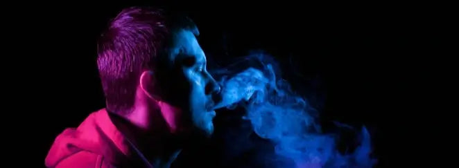 Person exhaling smoke against a dark background with a focus on the smoke and person's face.